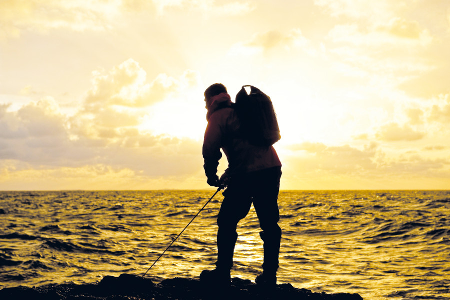 A man fishing on a rock looking out to sea and a sunset.